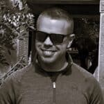 Man wearing sunglasses smiling outdoors in black and white.