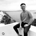 Man smiling, sitting on a white wall overlooking a coastal landscape.