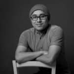 Young man with glasses and beanie, smiling, sitting on a chair.
