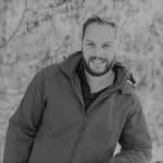 Man with beard smiling outdoors, wearing a jacket.