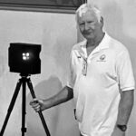 Older man with white hair holding a laser scanner on tripod.