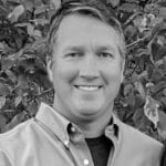 Smiling man outdoors with foliage in background.