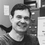Smiling man in an office environment with filing cabinets and shelves.