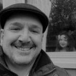 Man smiling with a young girl in a window behind him, rainy day scene.