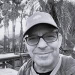 Man wearing glasses and cap outdoors with trees in background.