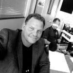 Man smiling at microphone in recording studio with another person in background.