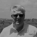 Man wearing sunglasses and a white shirt outdoors in a rural area.