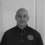 Man in black shirt with logo, standing indoors against white panel wall.