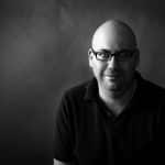 Black and white portrait of a man wearing glasses and a dark shirt.