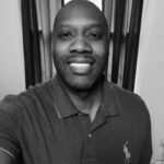 Portrait of Jay R. smiling, wearing a polo shirt, in front of curtains.