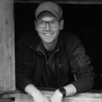 Man with glasses and cap smiling, leaning on wooden structure.