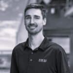 Smiling man in black polo shirt with logo, outdoor background.