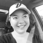 Woman smiling in car wearing a baseball cap and seatbelt.