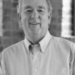 Portrait of a smiling man in a white shirt in front of a brick wall.