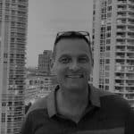 Man smiling on balcony with city buildings in background.