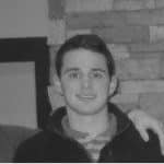 Young man smiling, casual attire, indoor setting with brick wall background.