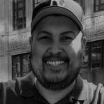 Portrait of Rudolph B. smiling, wearing a cap, in front of a building.