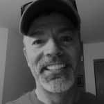 Black and white photo of a man smiling, wearing a cap, indoors.
