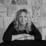Sherry Rennick smiling, sitting at a table with a stone wall background.