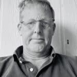 Man with glasses and short hair in front of wooden wall.
