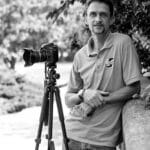 Thomas C. standing outdoors with a camera on a tripod near a river.