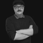 Portrait of Thomas W. with crossed arms, wearing glasses and a flat cap.
