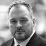 Close-up of a man with short hair and beard, wearing a suit, winking at the camera.