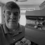 Man with mustache and polo shirt next to 3D laser scanner equipment.
