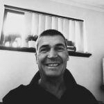 Smiling man with short dark hair in front of window with blinds.