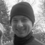 Smiling man wearing a beanie and neck gaiter outdoors in winter.