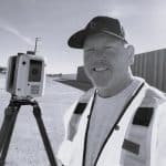 Man with cap and vest operating 3D laser scanner outdoors.