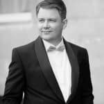 Man in tuxedo looking to the side, smiling, outdoor setting.