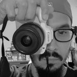 Man with glasses and cap taking photo with Canon camera in black and white.