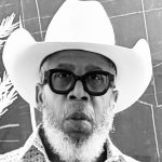 Man with white cowboy hat, glasses, and beard in front of a chalkboard.