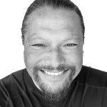 Smiling man with beard and slicked-back hair in black shirt.