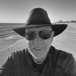 Man wearing a hat and sunglasses at the beach, smiling for a selfie.