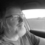 Man with glasses and beard driving in a car, looking out the window.