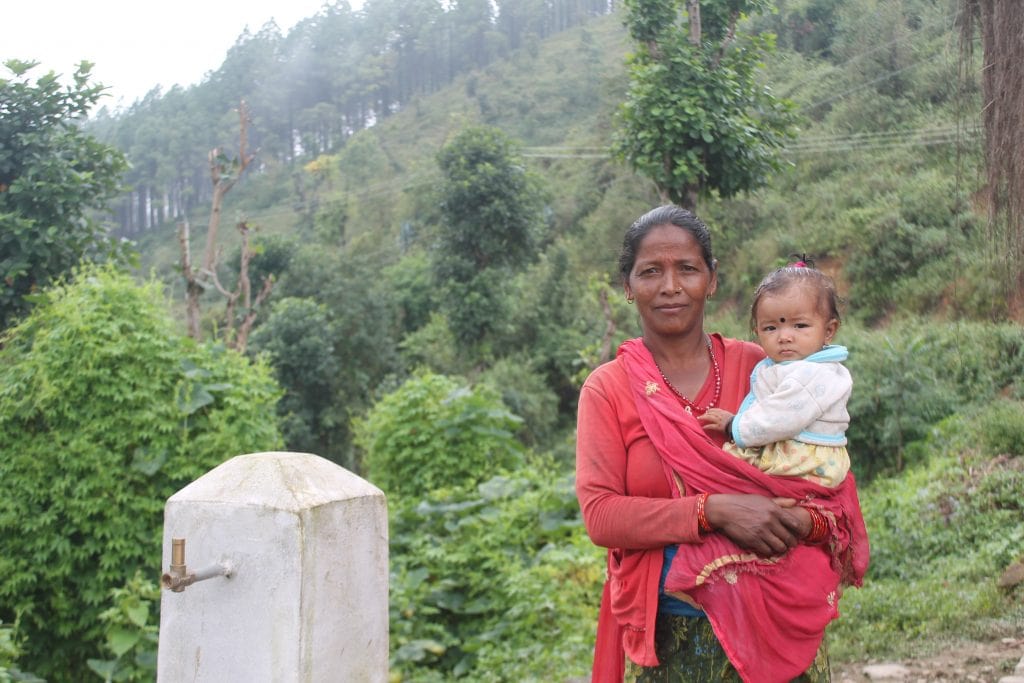 Chandra Kauli Sunar, A Nepali lady standing next to a water tap holding he young granddaughter