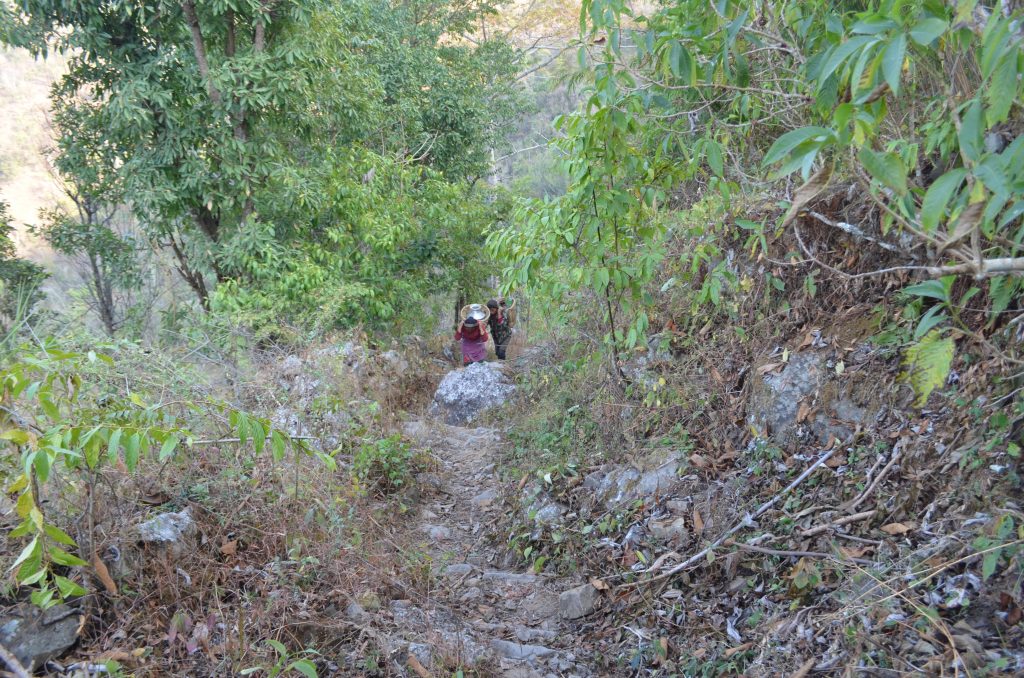 Looking down a steep hill path with two women climbing up carrying heavy loads of water on their backs