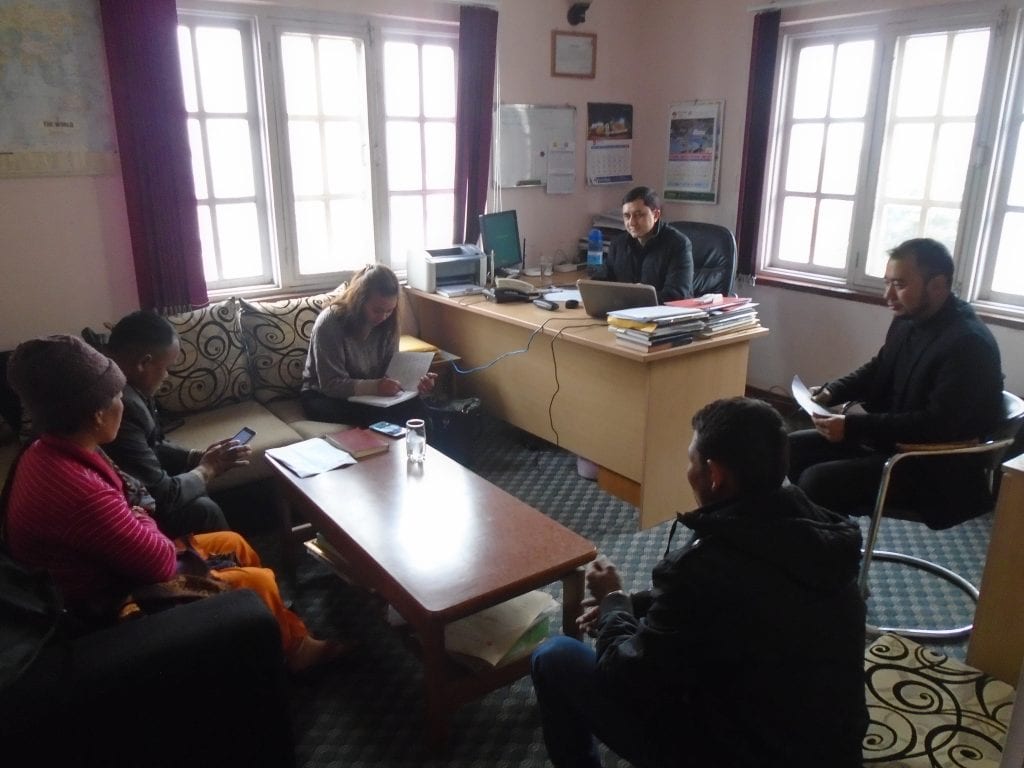 The photo shows a meeting between  Renewable World representatives, community representatives, and the Local NGO. Six individuals sitting in an office, one lady is taking ones. 