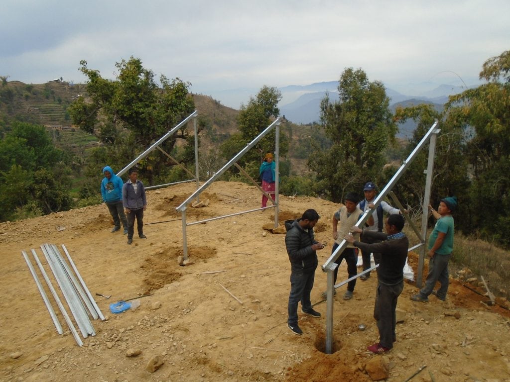 A metal frame, consisting of three triangular pieces, being erected on the hillside in Nepal with a eight community members inspecting the work 