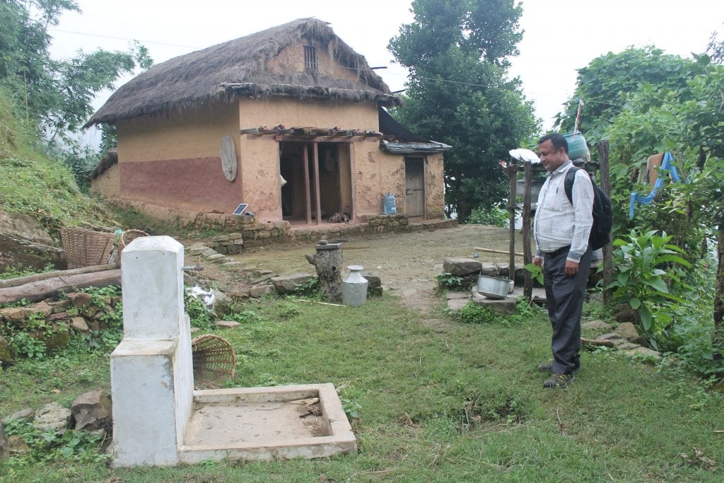 A tap stand built in the traditional Nepali style, painted while, in front of a traditional Nepali house. 