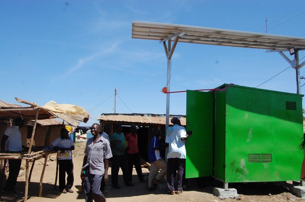 Photo of a solar microgrid. A large metal container box painted green with a structure above it supporting solar panels. Community members are milling around 