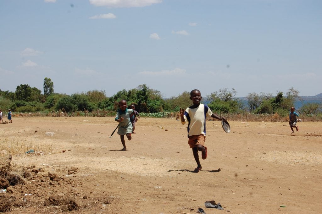 Four smiling children running towards the camera. The ground is dry and dusty, the sky is blue and clear 