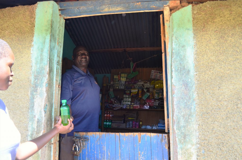 Pascal standing in his store serving a customer. Biscuits, crisps, soap and other items for sale can be seen behind him. 