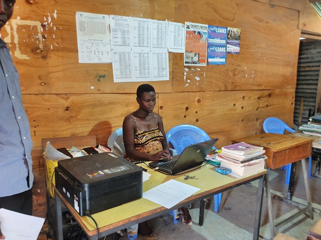 A teacher sitting at a desk using a laptop. A printer is on the table to her right, printed information is pinned to the wall behind. Preparing coursework and exams is now much easier for teachers, as they can power a computer and printer at the school