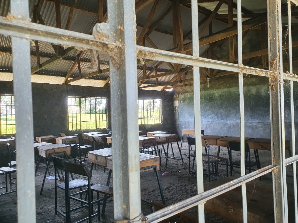 Looking through the window of a classroom at Markmatunga Secondary School on Kiwa Island. A bare room with basic chairs and tables, some broken, a dusty floor, a large chalk board on the wall, and two electric lighbulbs fitted to the ceiling powered by the solar microgrid installed on Kiwa Island 