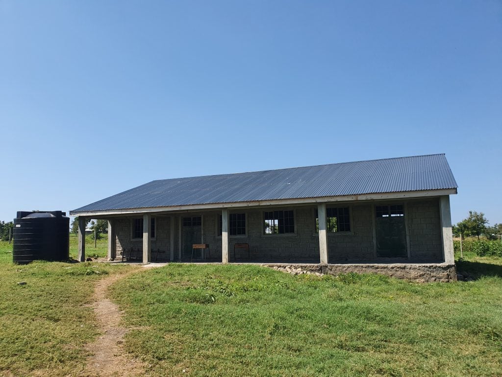 A very basic school building pictured from the outside. Bright blue sky overhead and a dirt path leading up to the classrooms. 