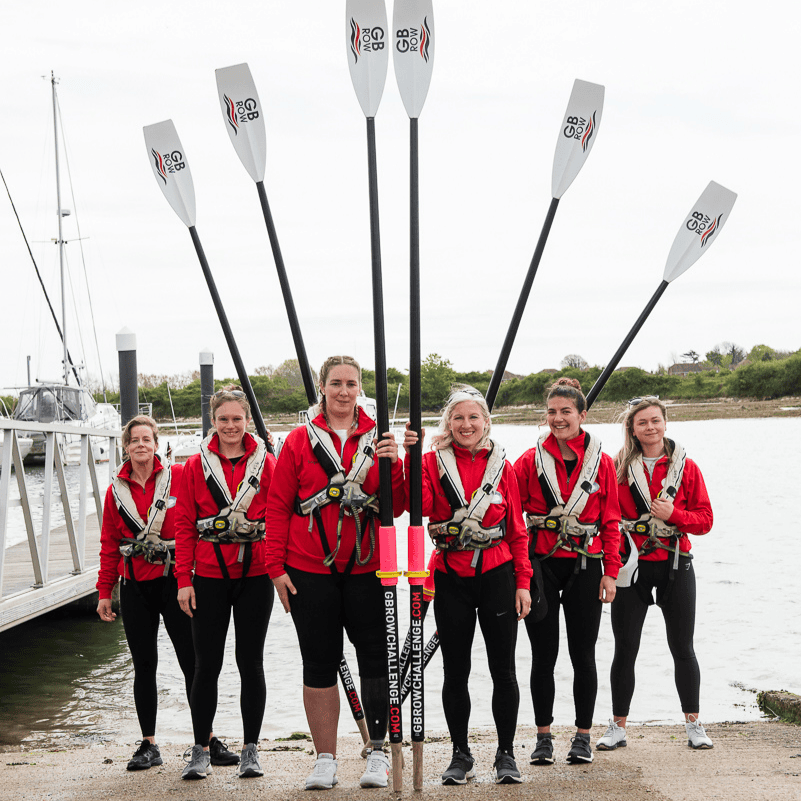 Six female rowers standing on the bank of a river, each woman is holding up a tall rowing oar that towers above their heads.