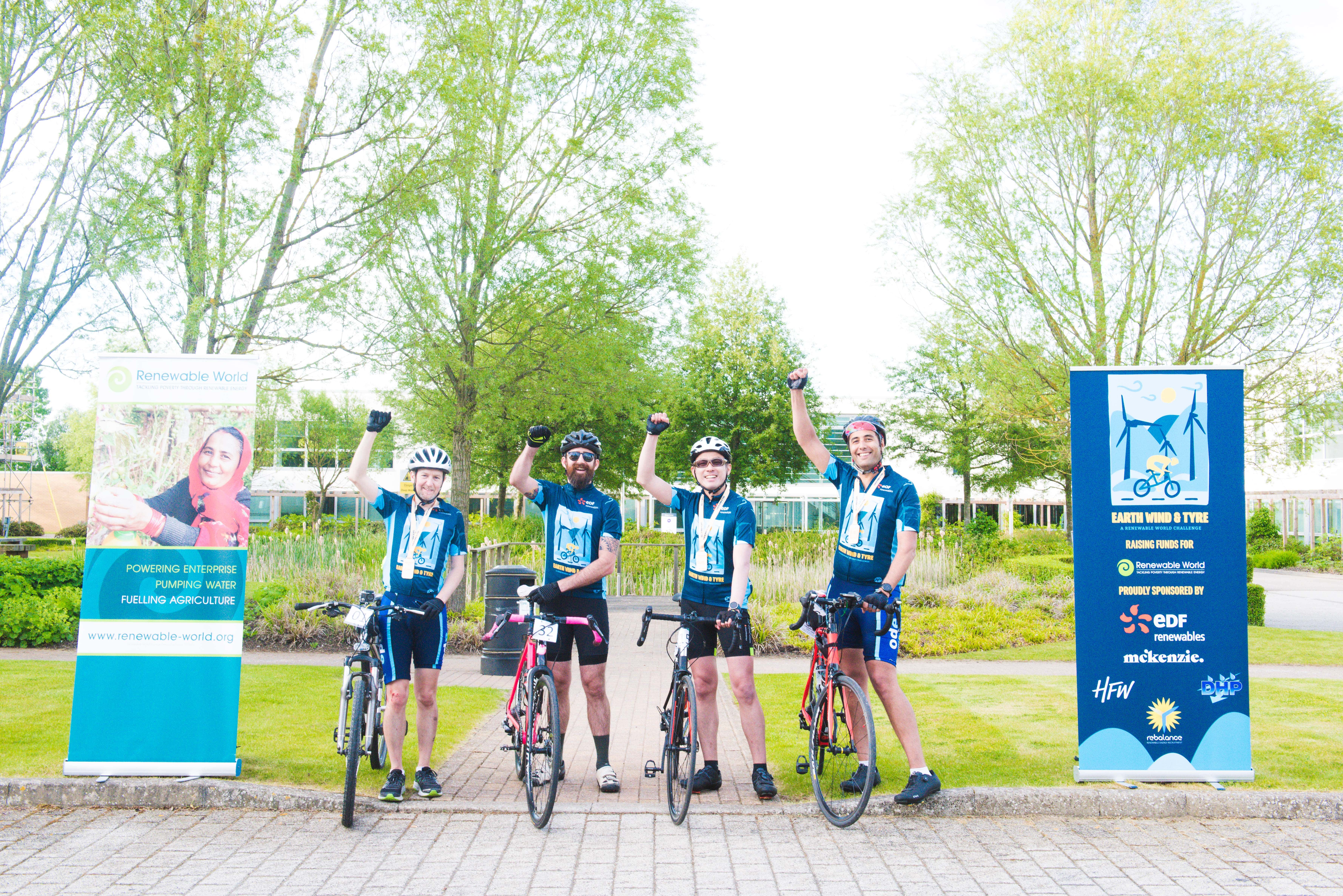 Four cyclists are pictured at the finish line with their bikes in between two event banners, they each have an arm raised in the air in celebration of their achievement